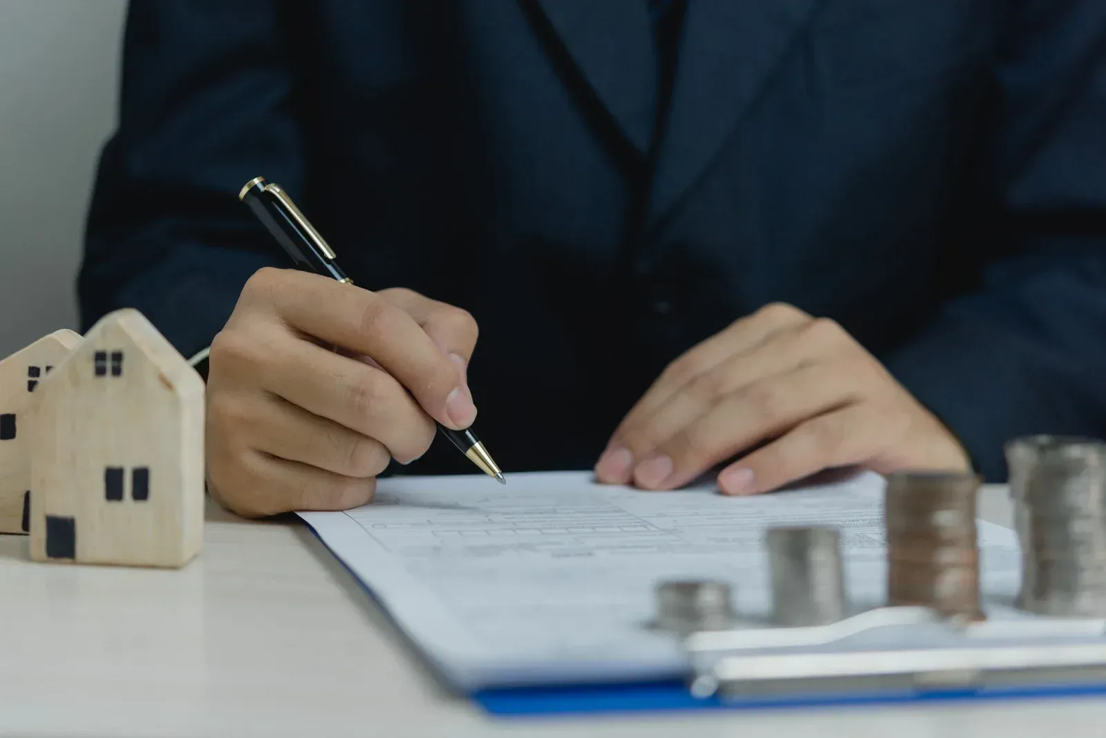 Person signing document with pen, miniature houses, and stacks of coins on a table.