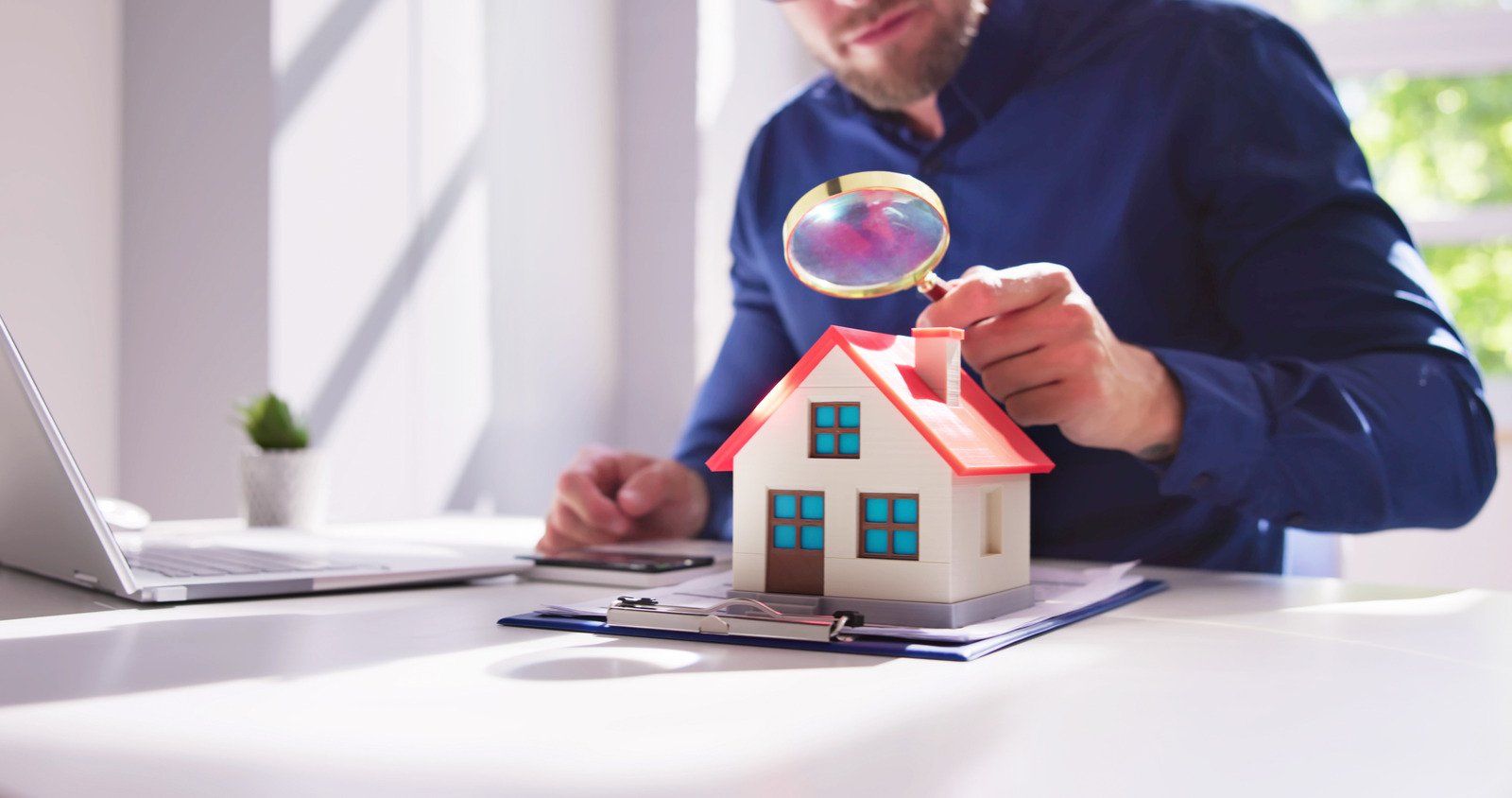 Person examining model house with magnifying glass on a desk with laptop and paperwork.