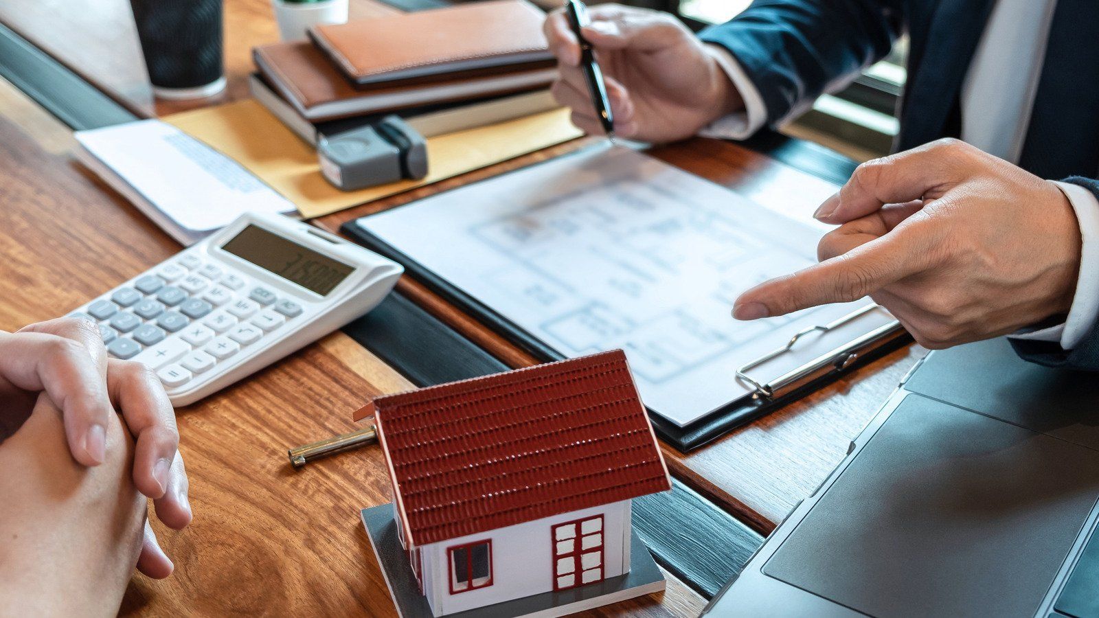 Real estate agent pointing at paperwork, discussing terms with a client; miniature house on table.