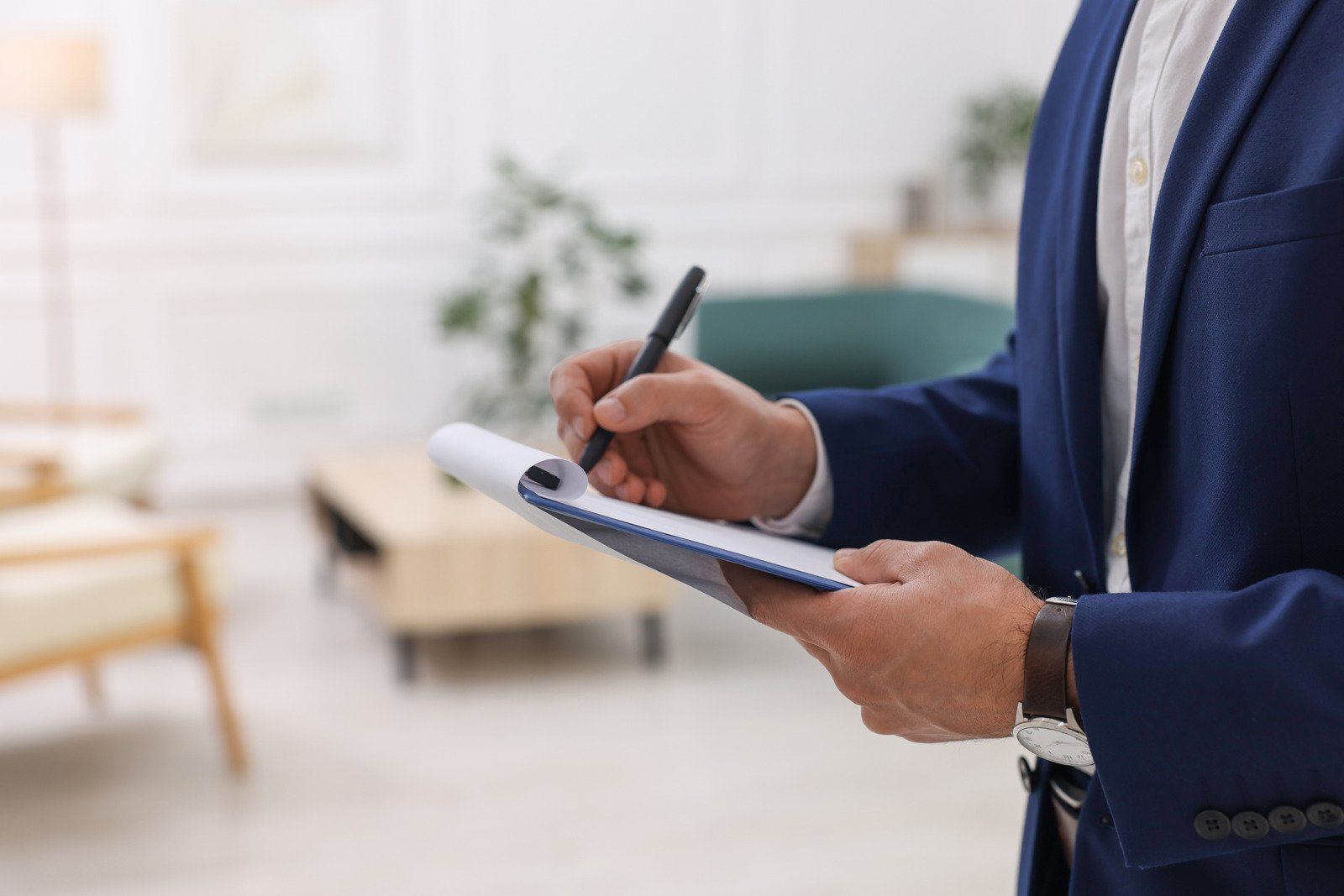 Man in a blue suit writes on a notepad inside a living room.