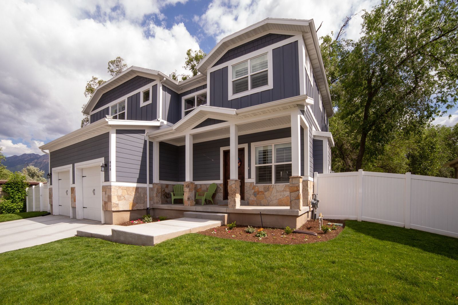 Two-story blue house with porch, tan accents, and two-car garage, set on green lawn with white fence.