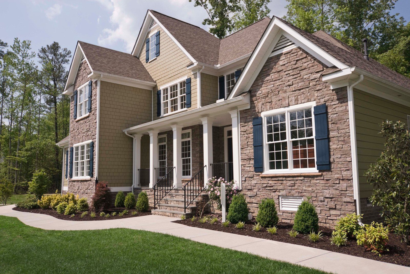 Two-story house with tan siding and brick facade. Porch with white columns, blue shutters, and green lawn.