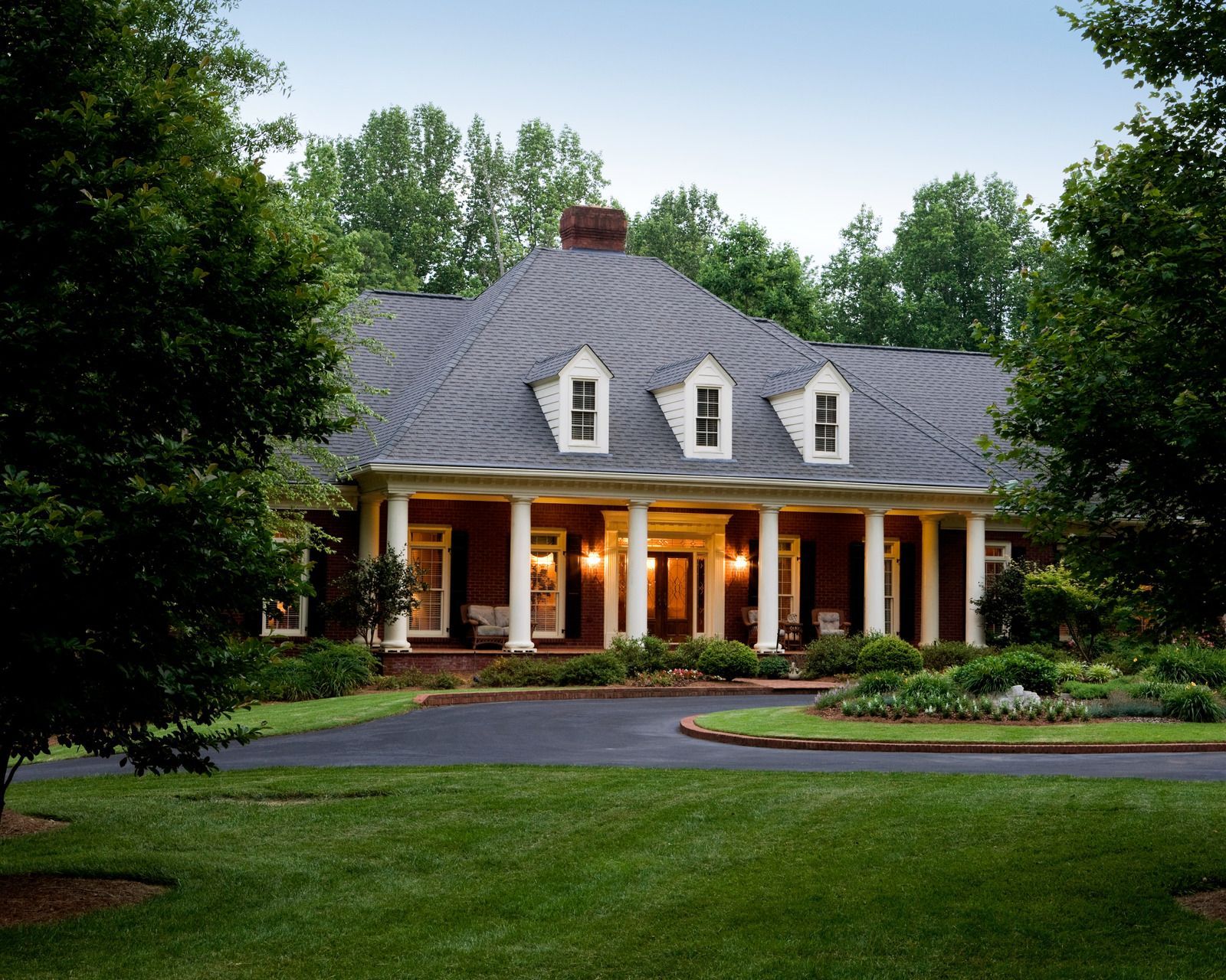 Elegant brick home with white columns, a long porch, and a gray roof, set among trees and green lawns.