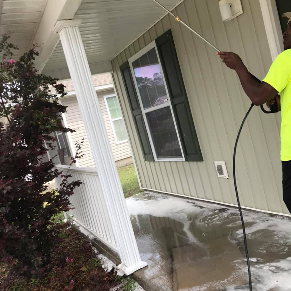Man power washing a porch with a pressure washer; the man is wearing a neon yellow shirt.