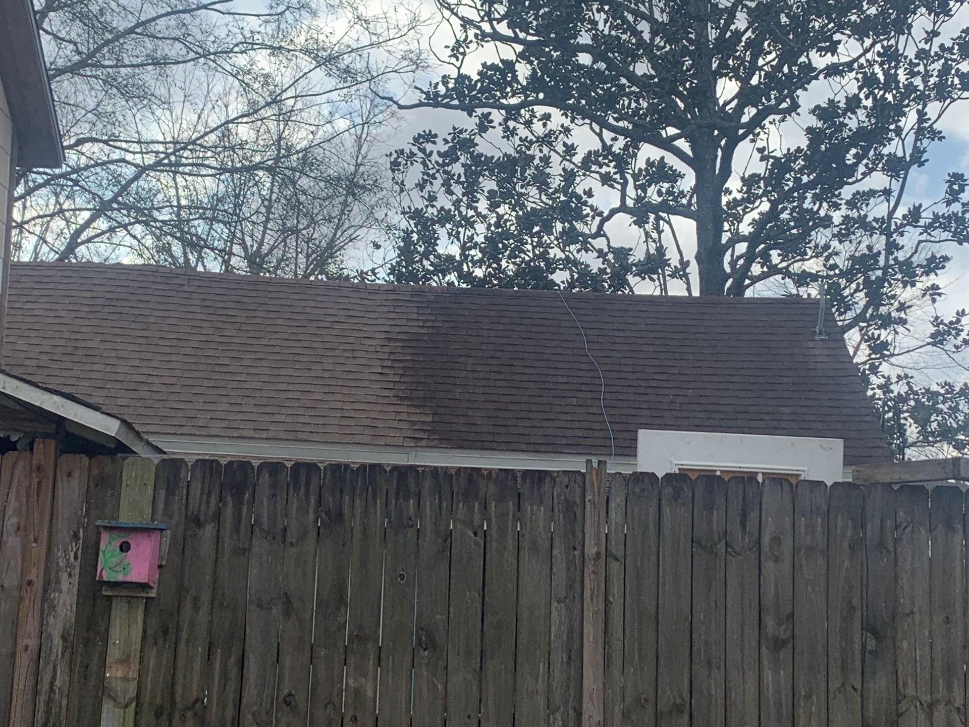 Brown roof of a building behind a wooden fence. A birdhouse hangs on a fence post. Trees in background.