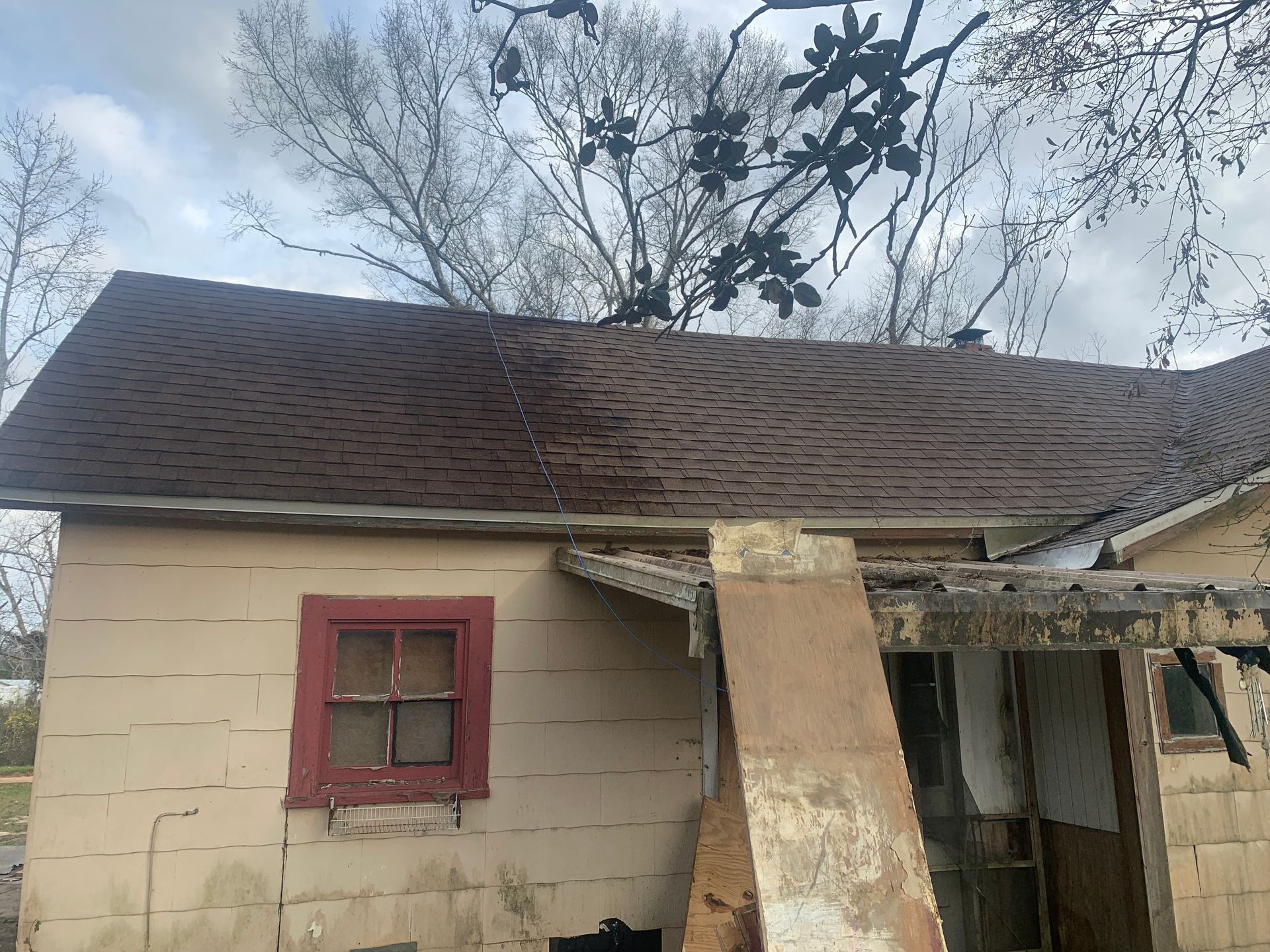 Old house with brown roof, tan siding, red window frame, and damaged porch.