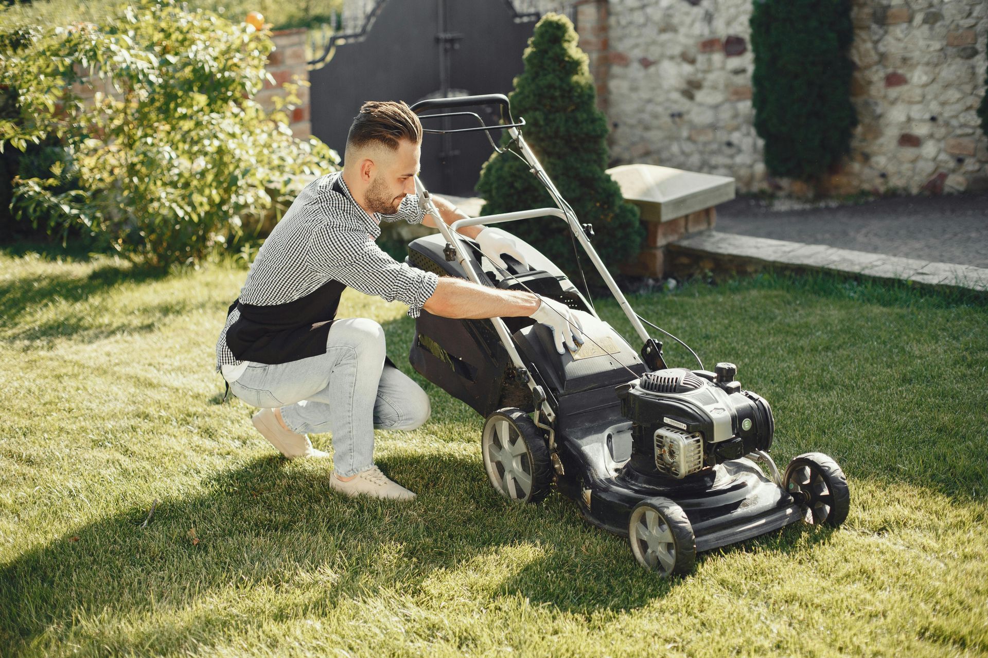 Man kneels to adjust a lawnmower on a grassy lawn in a sunny backyard.