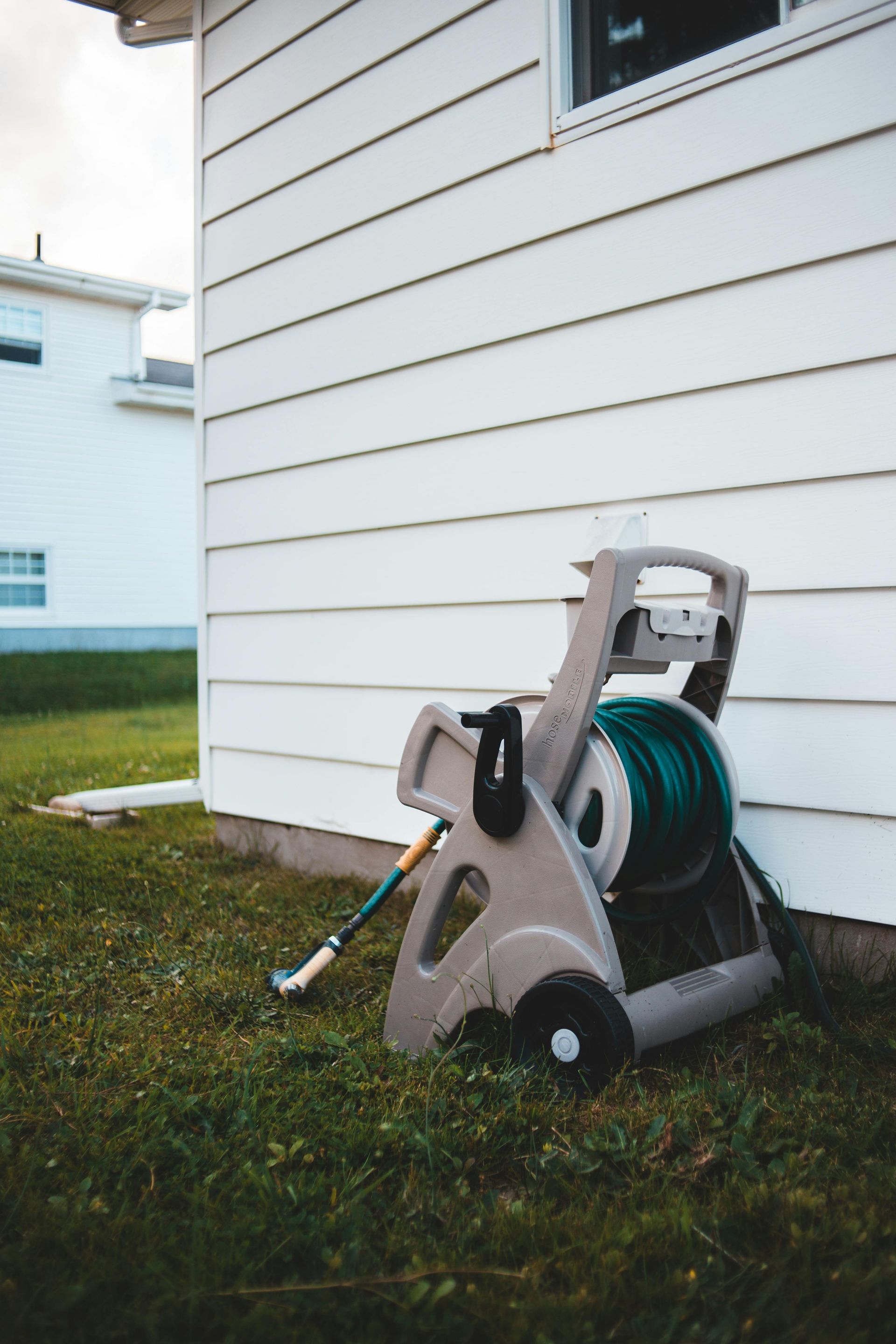 Gray hose reel with green hose by a white house on a grassy lawn.