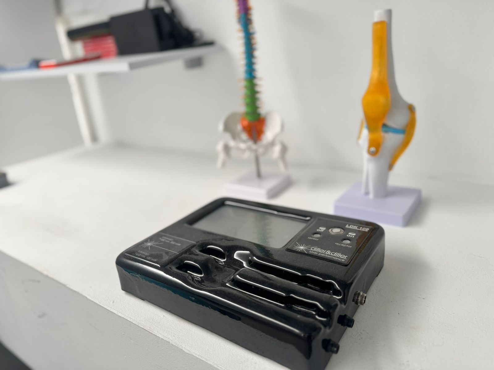 Unidad TENS negra sobre una superficie blanca, con modelos de columna y rodilla en el fondo.