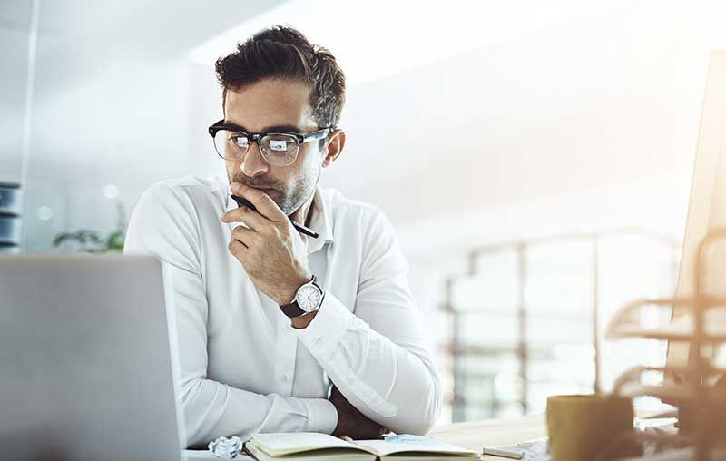 A man wearing glasses is sitting at a desk in front of a laptop computer.