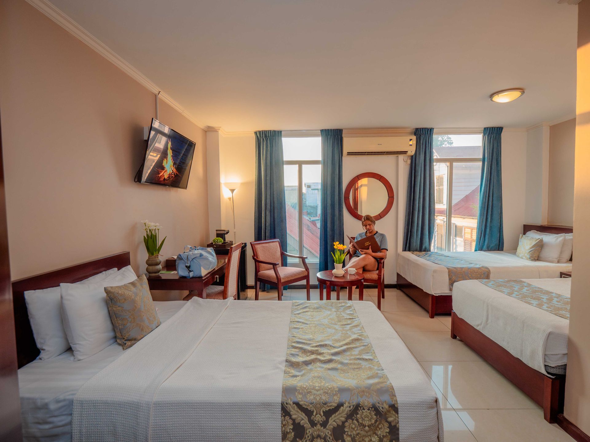 Hotel room with three beds, chair, table, and a person sitting near the window.
