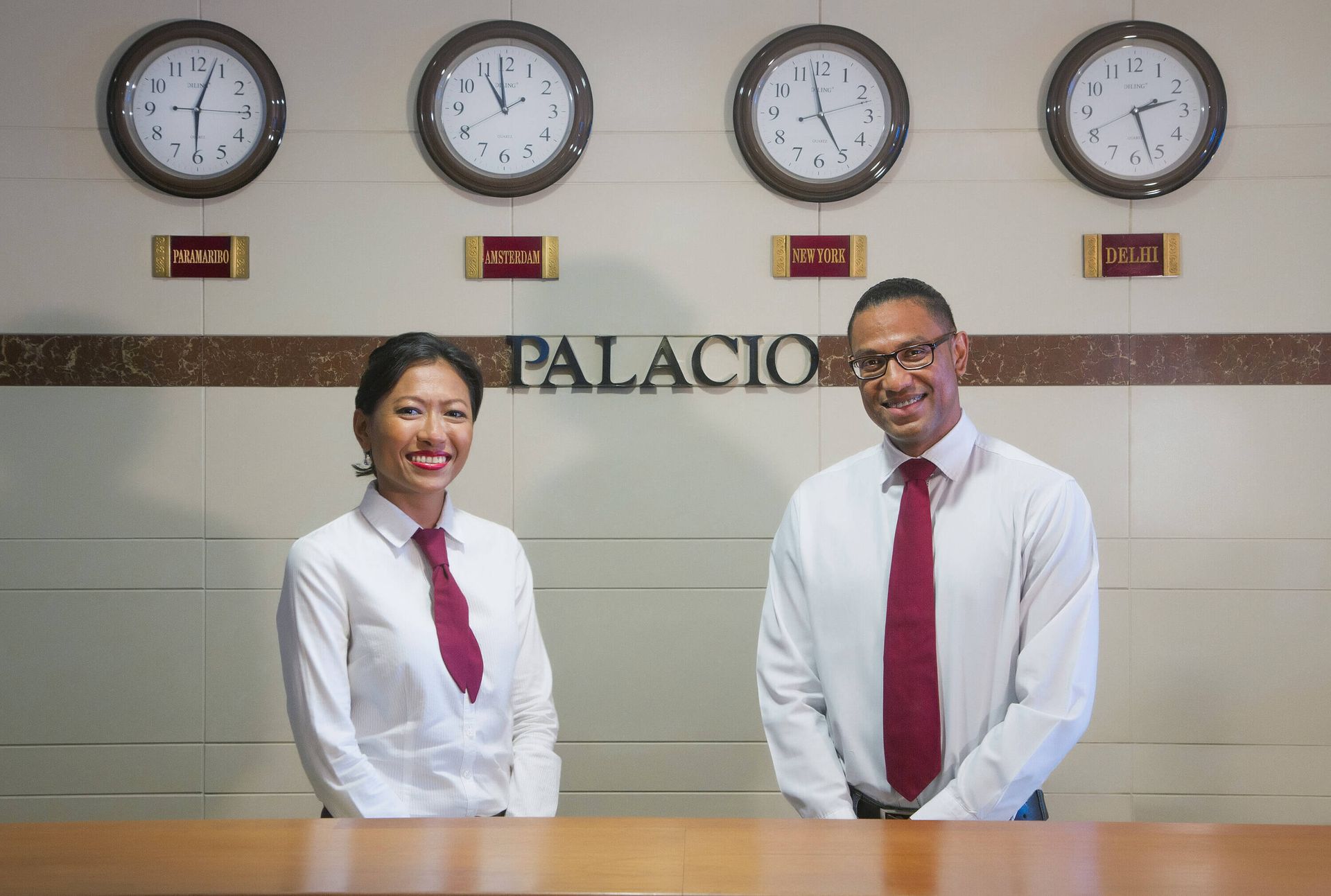 Two hotel receptionists behind a counter, smiling at the camera. Beige wall with clocks showing different time zones,