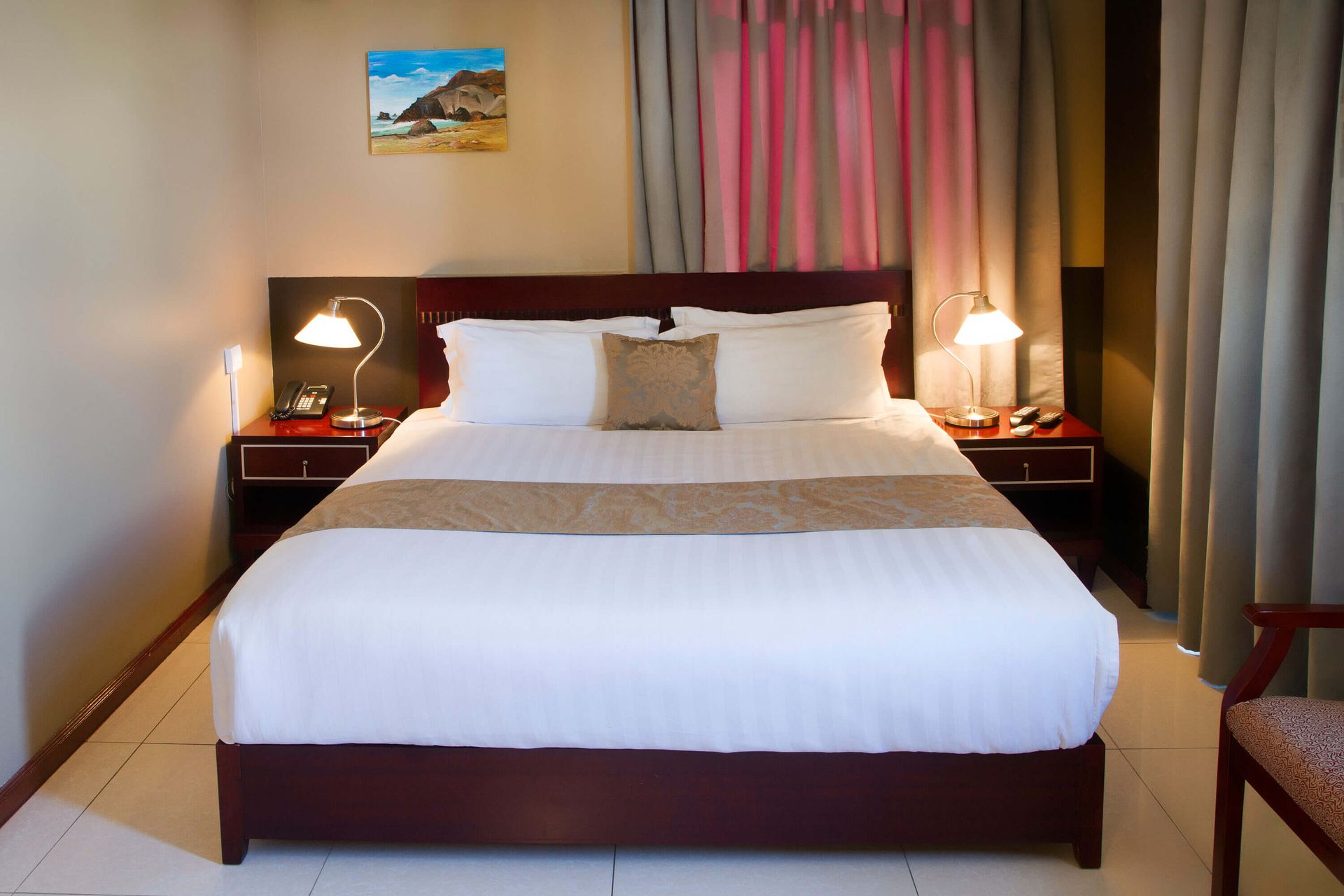 Hotel room with a bed, bedside tables, and a small painting on the wall. The room is in warm tones.