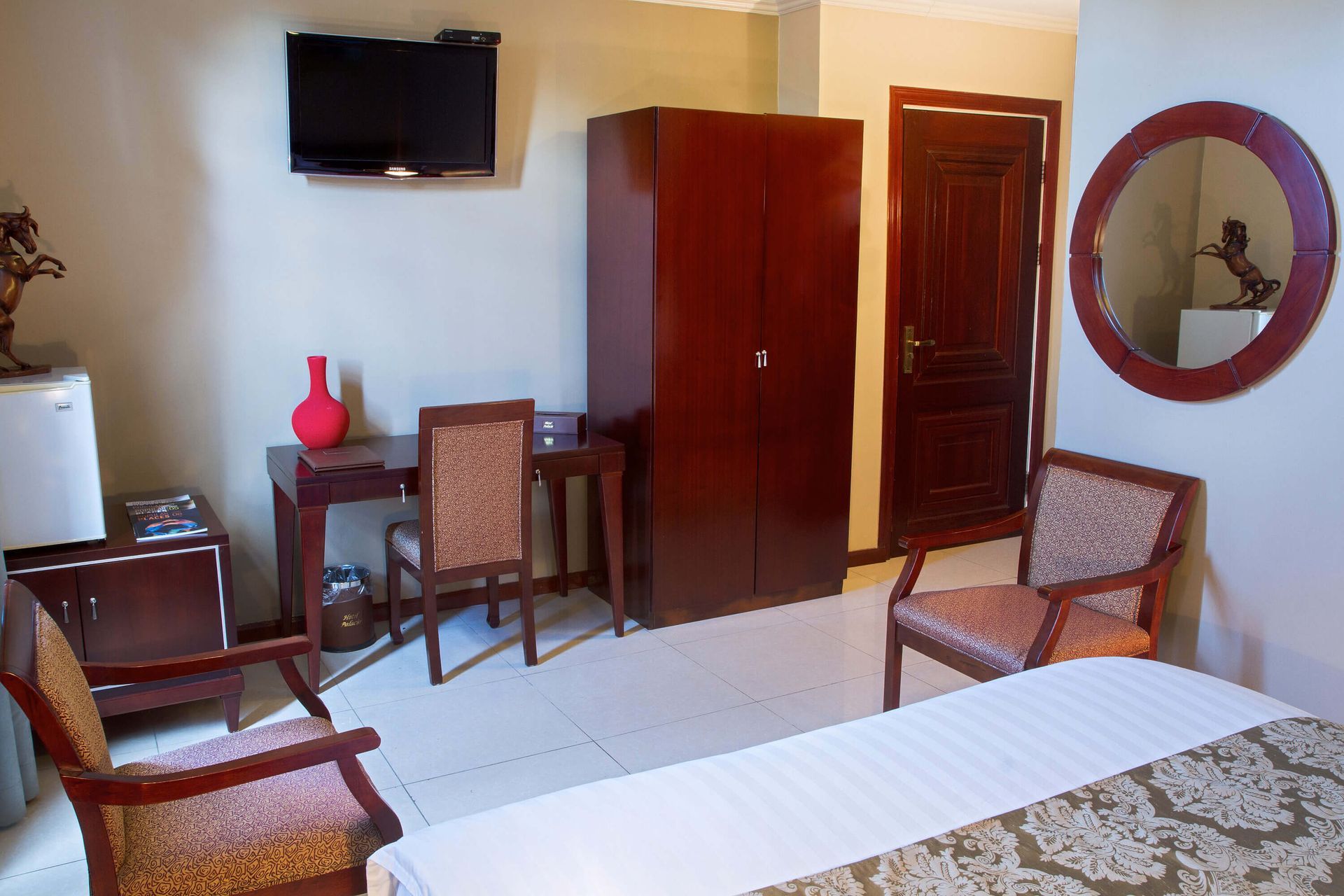 A hotel room with a bed, desk, wardrobe, television, chairs, and a round mirror. Beige walls and white tiled floor.