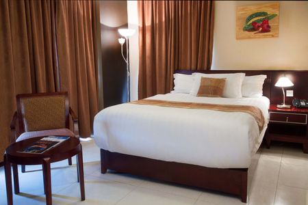 Hotel room with a king-sized bed, a small table with a chair, and brown curtains. A painting hangs on the wall above the bed.