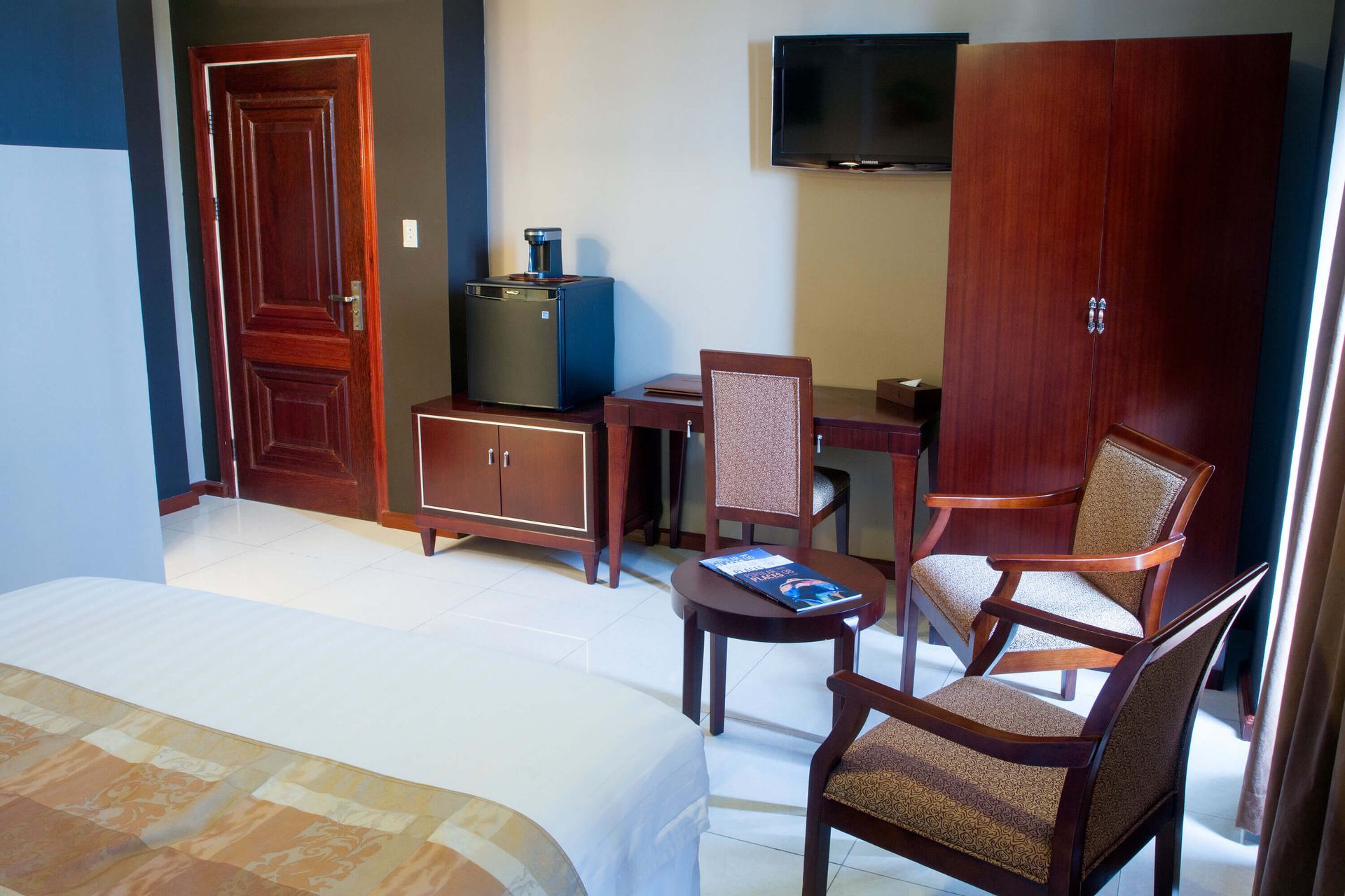 Hotel room with a bed in the foreground, a wooden door, cabinet, desk, chairs, and television.
