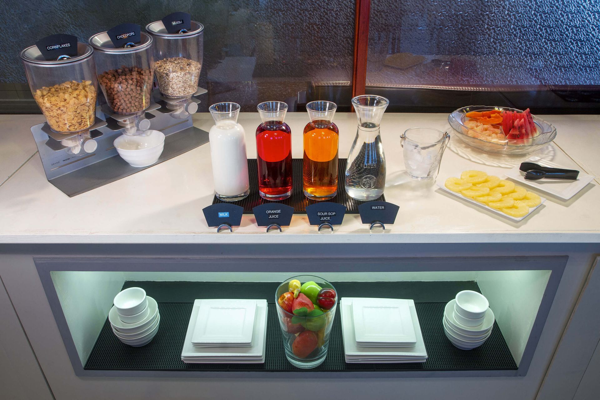 Breakfast buffet with cereal dispensers, juice carafes, fruit, and dishes. A bright, well-lit setting.