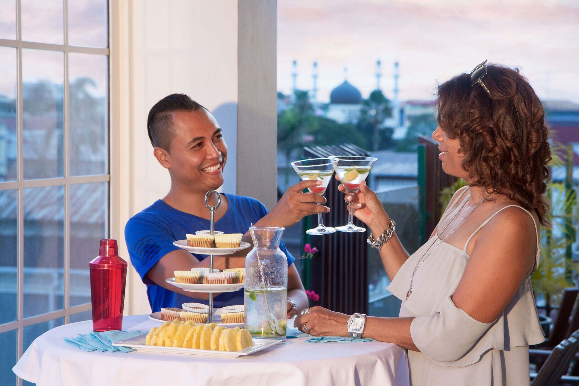 Two people clink martini glasses at a rooftop table, smiling. A tiered tray of desserts and a pitcher of water are on the table.