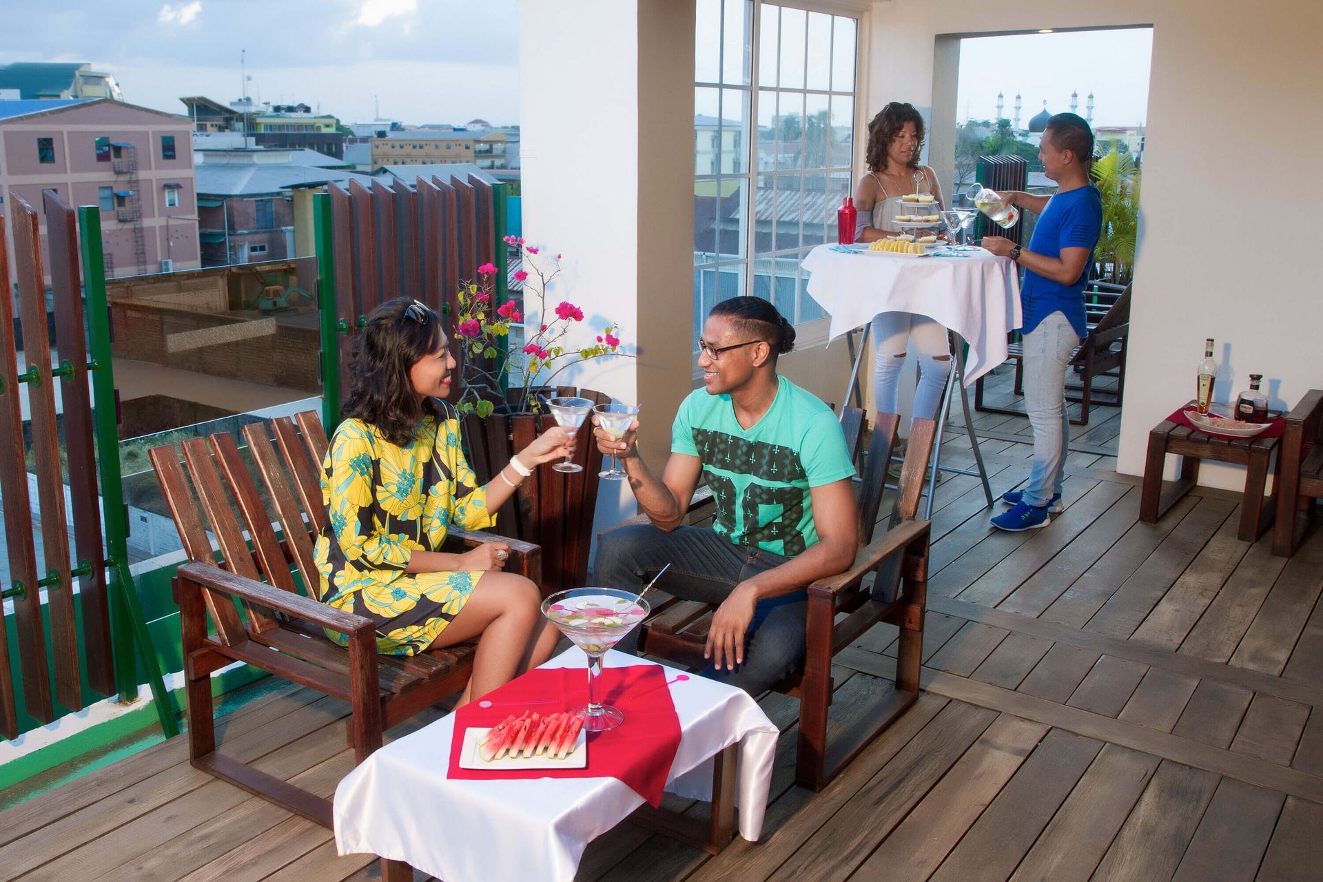 People on a rooftop patio clink glasses. Two people are seated at a table, while others stand near a bar table with drinks, overlooking a city.