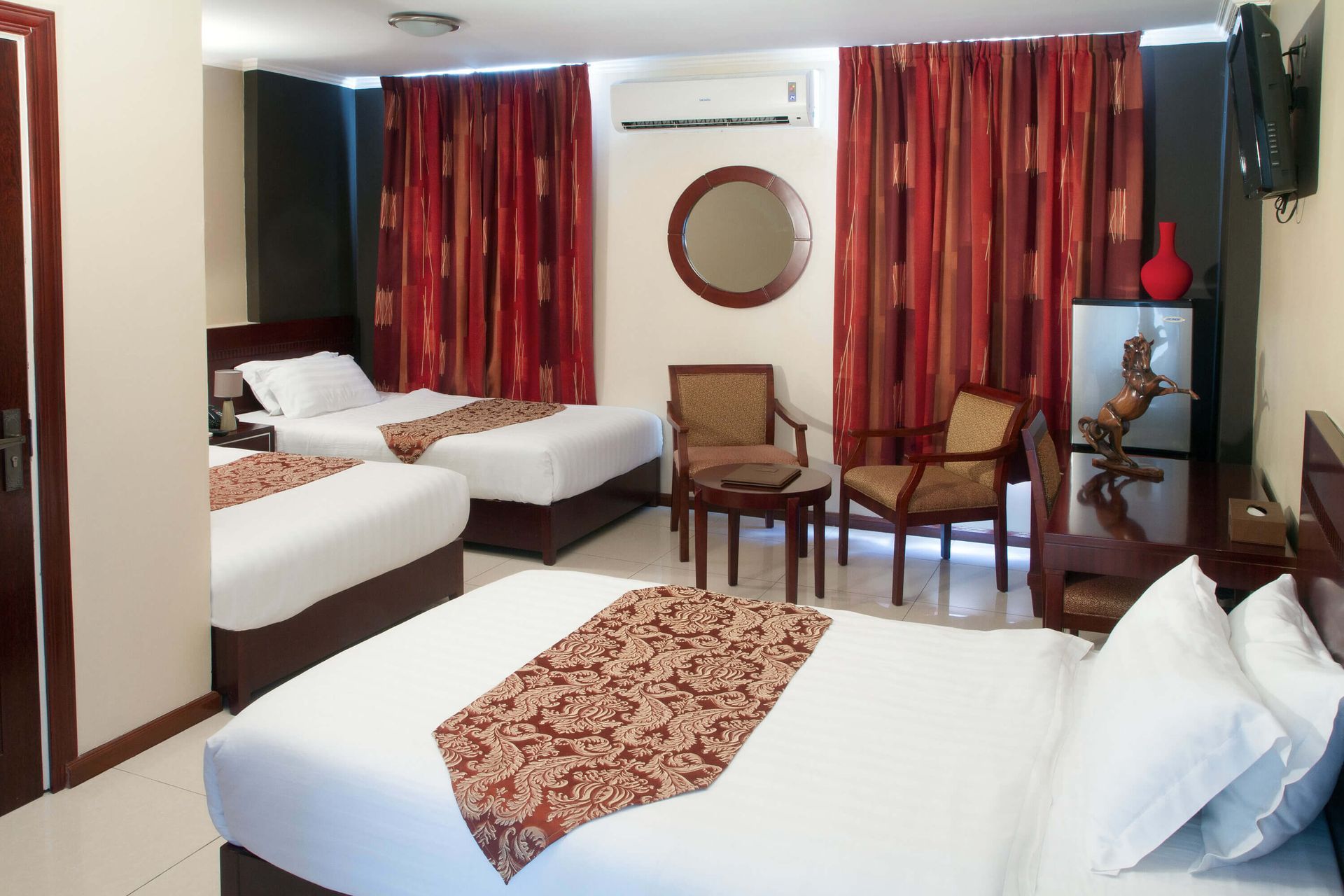A hotel room with three beds, red curtains, a mirror, and two chairs.