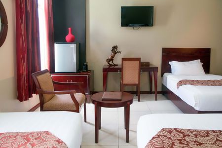 A hotel room with two beds, a mini-fridge, a desk, and a TV. Furniture is dark wood with red and brown accents.