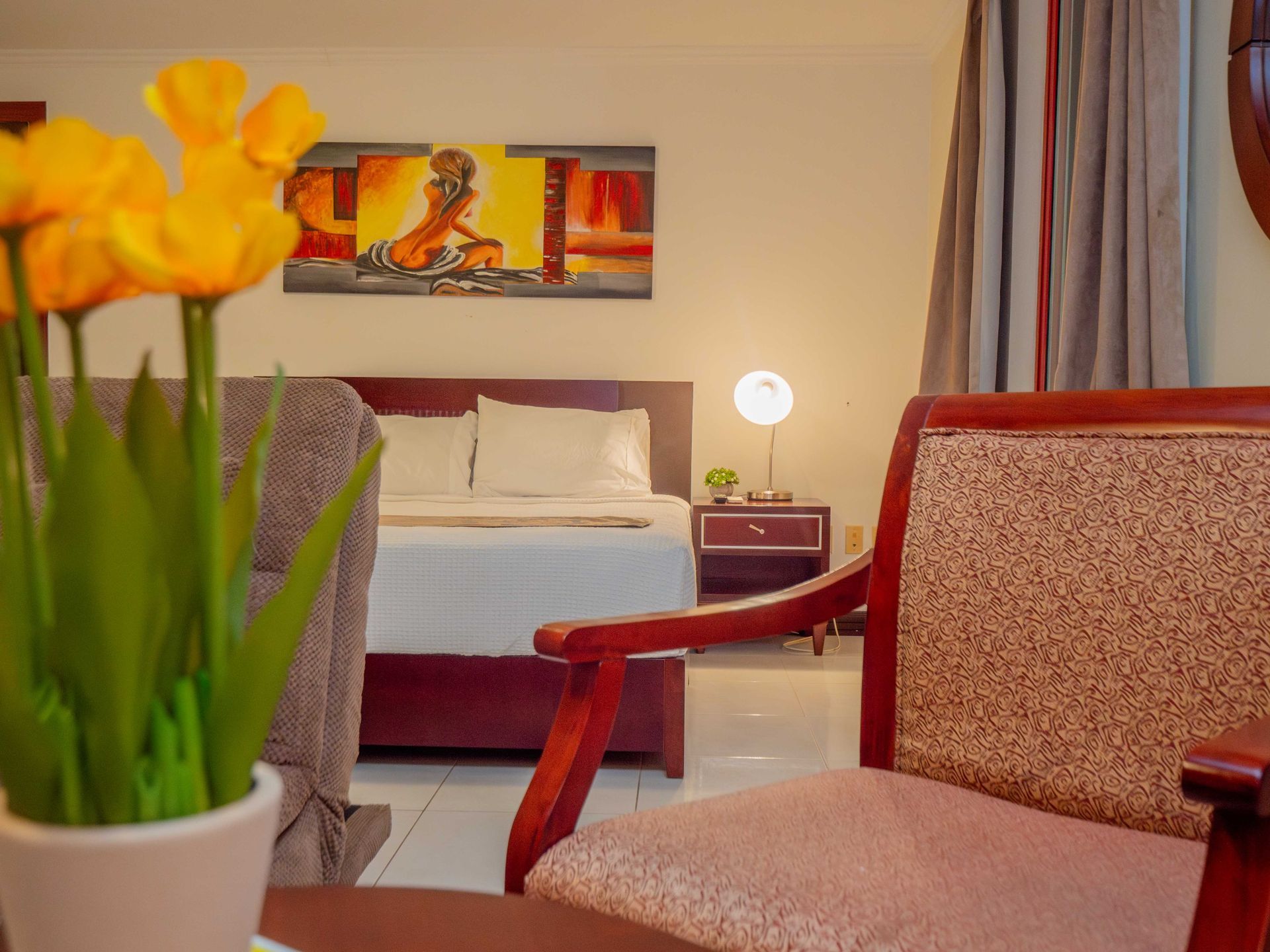 Bedroom with bed, chair, and art, featuring a vase of yellow flowers.