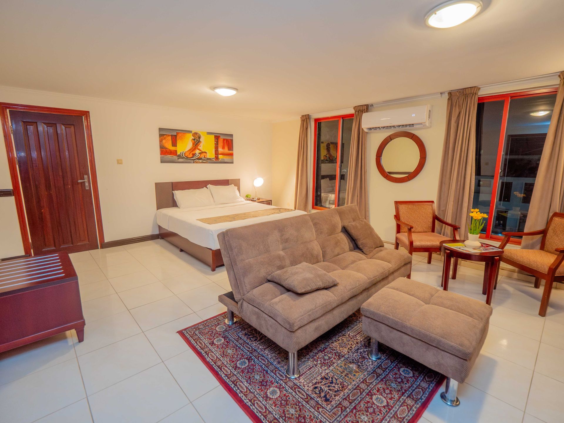 Hotel room: bed, sofa, chairs, rug. Neutral walls, red accents.