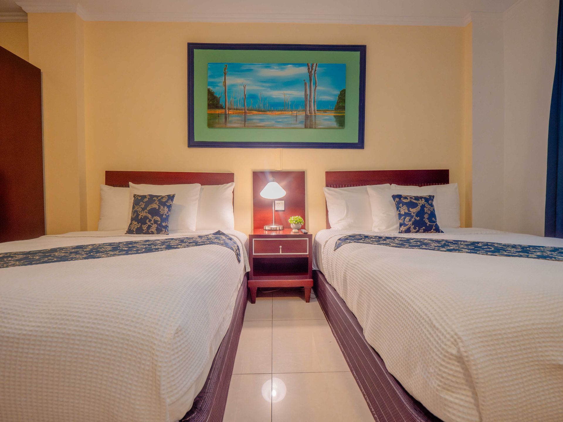 Two twin beds in a hotel room with a nightstand, artwork, and beige walls.