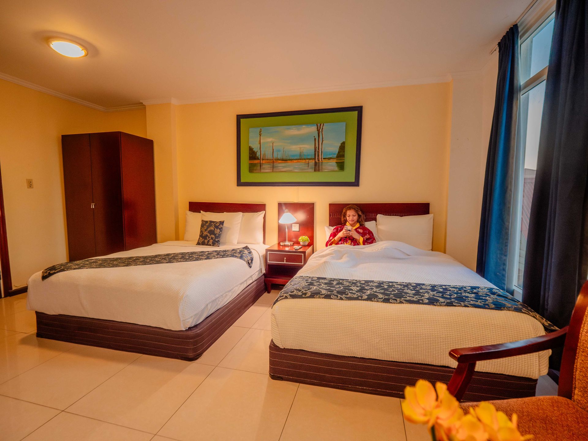 A hotel room with two twin beds, one occupied by a woman reading. Wooden furniture and artwork decorate the room.