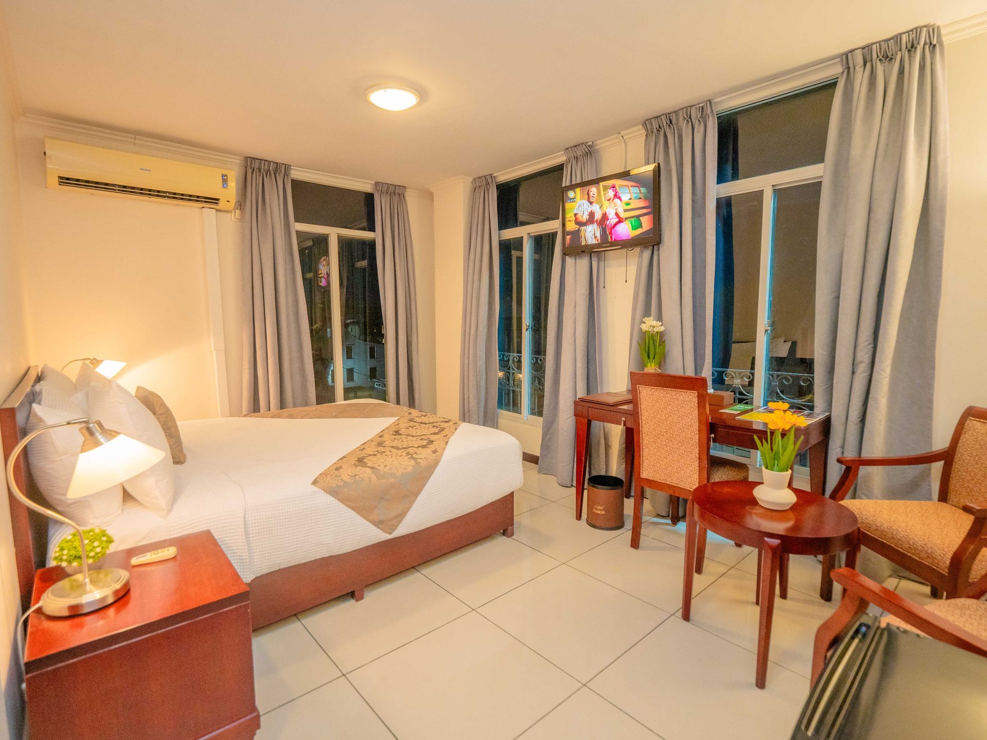 Hotel room with a bed, desk, chairs, and a view through windows.