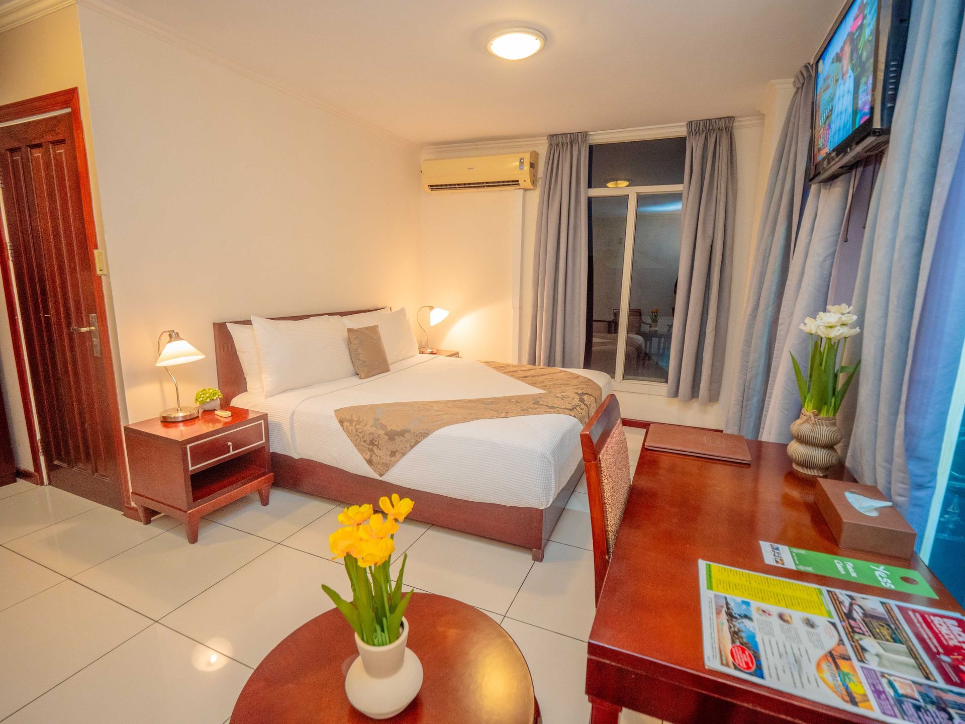 Bedroom with a double bed, desk, and wooden furniture, white walls, and natural light.