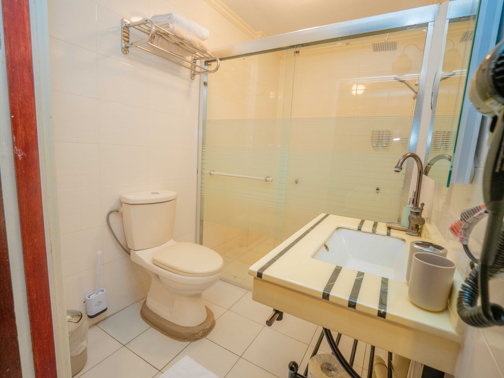 Small bathroom with toilet, shower, sink, and hair dryer. Beige walls, tiled floor.