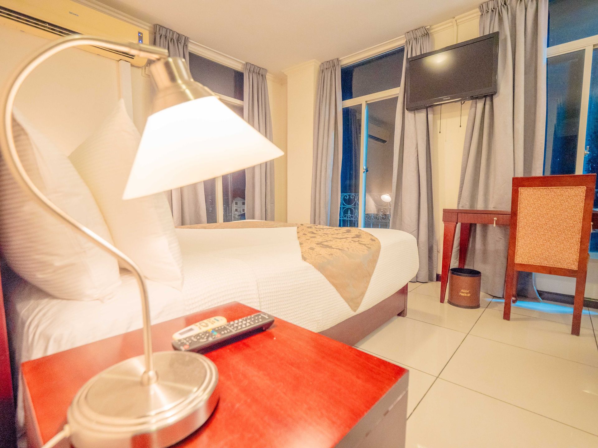 Hotel room with bed, desk, TV, and a lamp. White walls, light-colored linens, and a red bedside table.