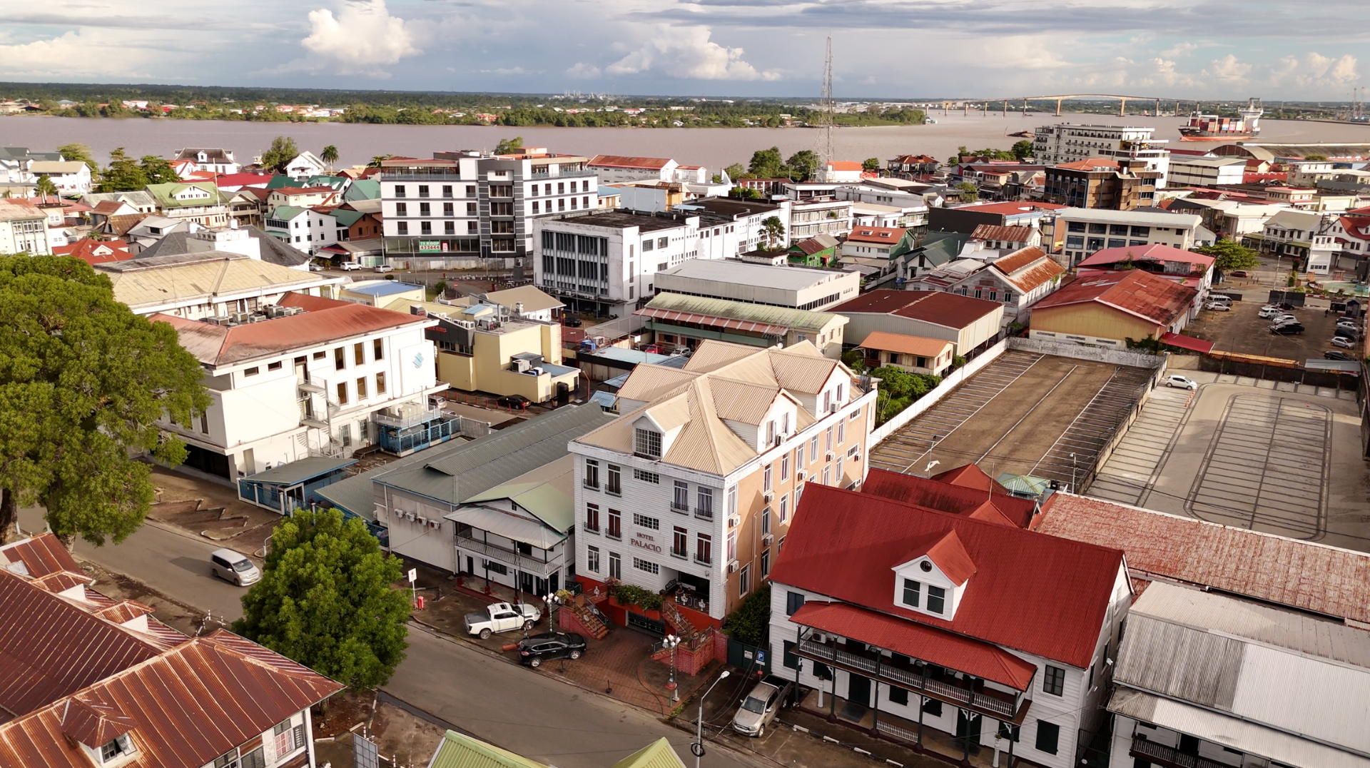 Guests checking in at Hotel Palacio’s wooden front desk in Paramaribo, Suriname — with clocks above and suitcases by their side.