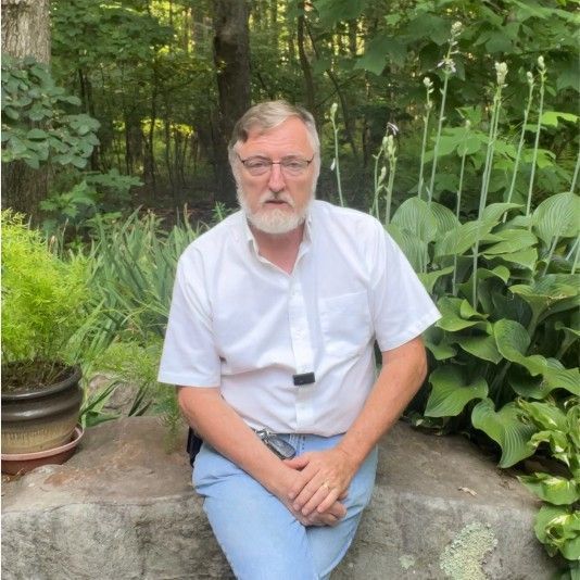 Man with a white shirt and glasses sits on a rock, hands clasped. Surrounded by plants in a garden.