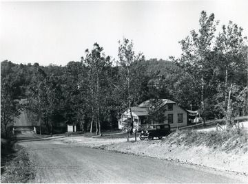 Dirt road leading to a two-story house surrounded by trees; a car is parked in the driveway.