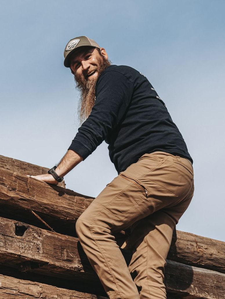 Man with a long beard climbing logs, smiling, wearing a baseball cap, dark shirt, and tan pants. Sky in the background.