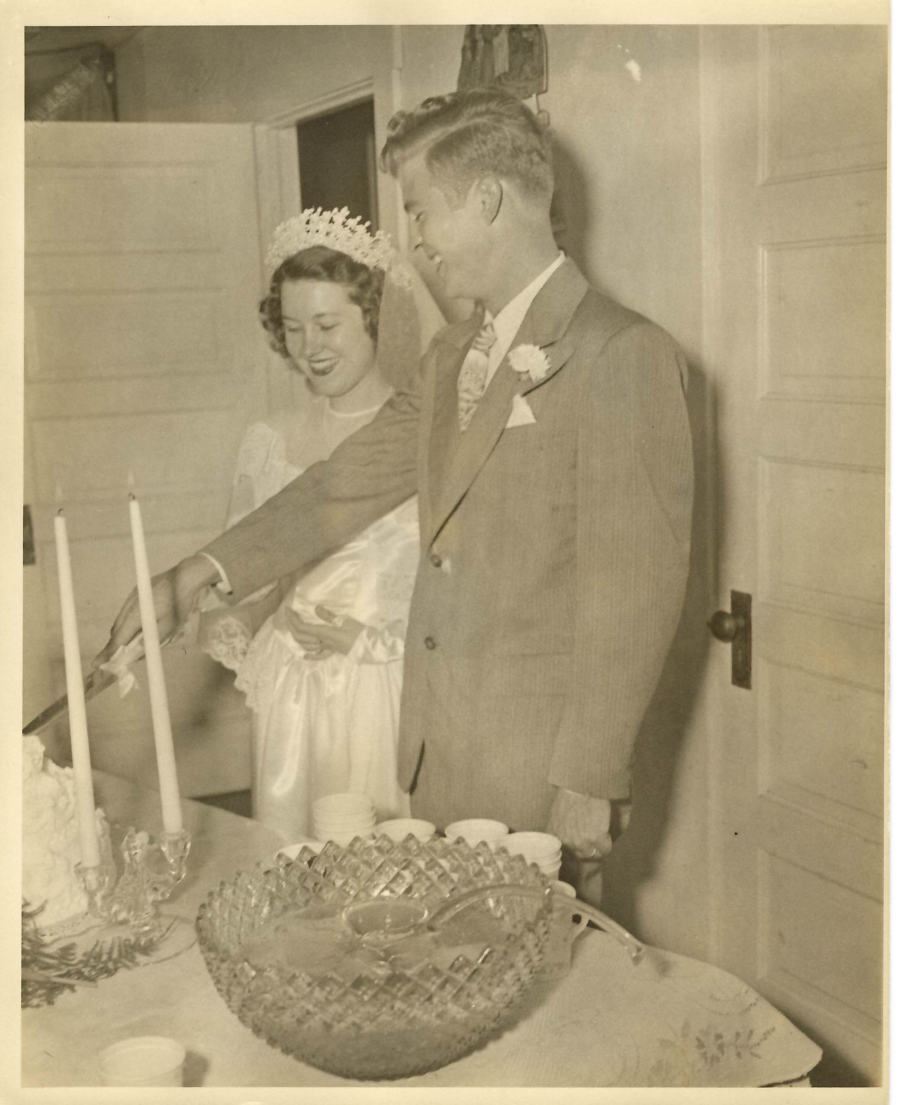 Bride and groom cutting wedding cake. Bride wears a tiara and gown. The groom wears a suit. Indoors.