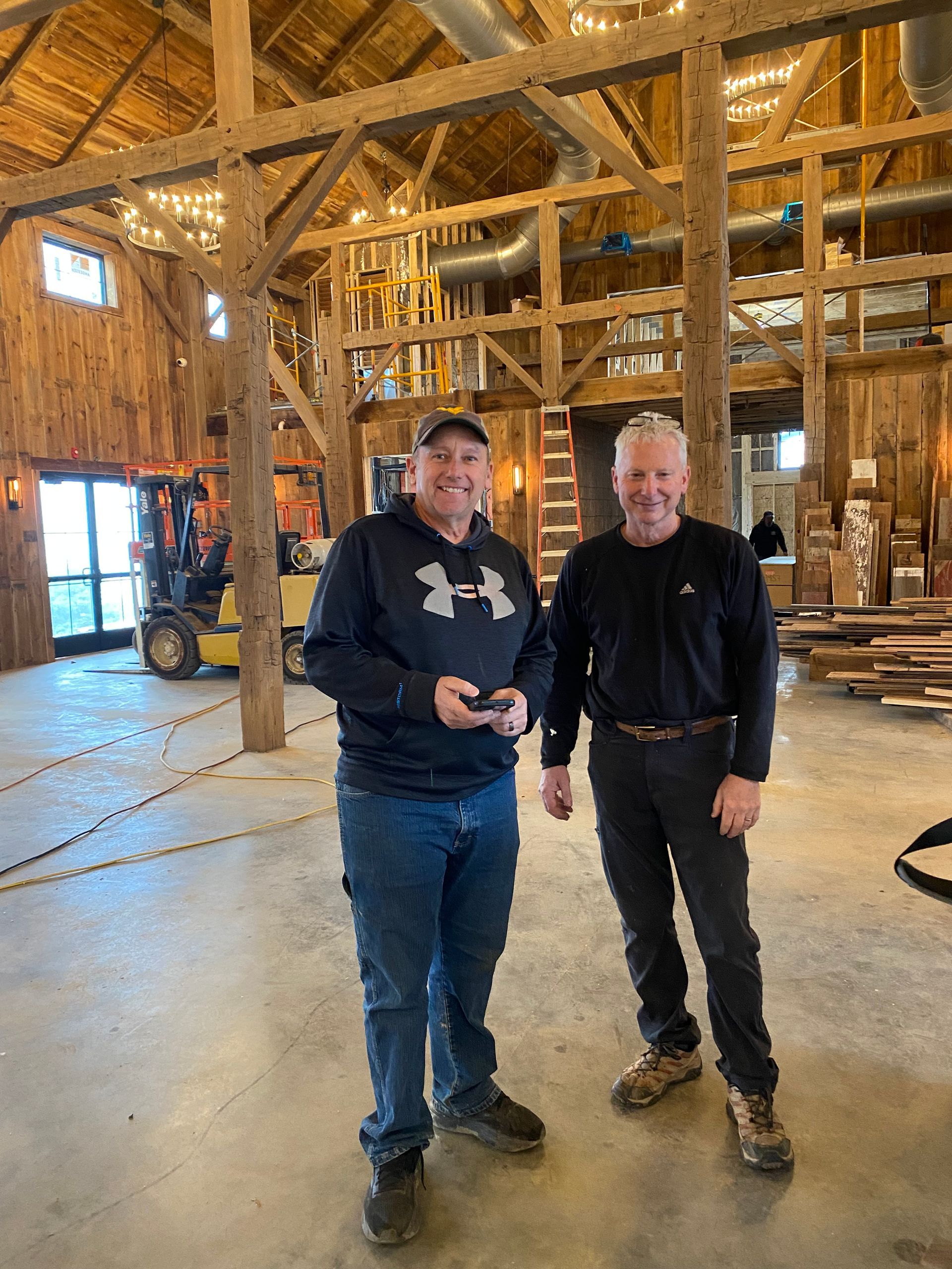 Two men stand inside a large wooden barn, one in a hoodie holding a phone, the other in a black shirt, both smiling.