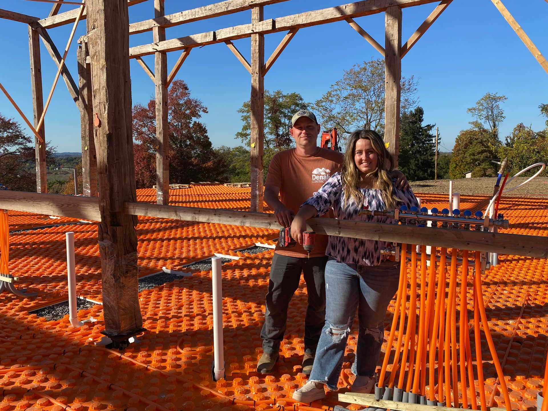Two people stand inside a barn frame. Orange flooring and structural beams are visible against a blue sky.