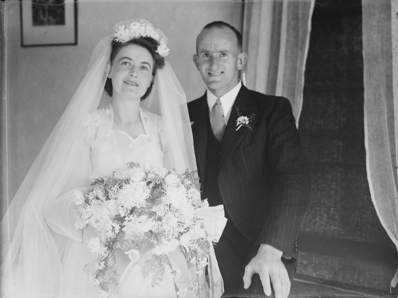 Bride and groom in wedding attire, holding bouquet. They stand near a doorway.