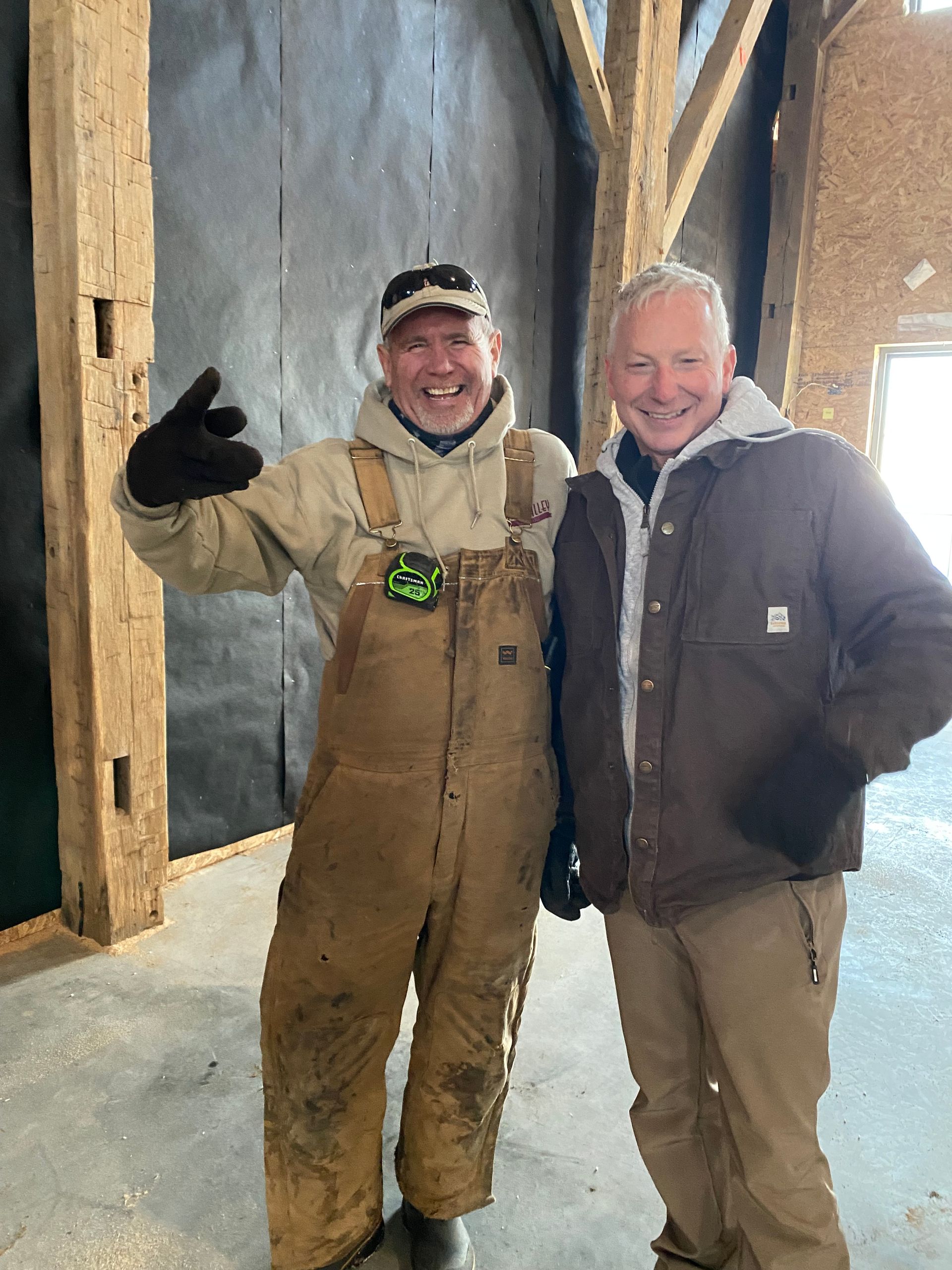 Two men pose in a barn. One in overalls gives a peace sign, the other smiles. Brown beams and dark wall.