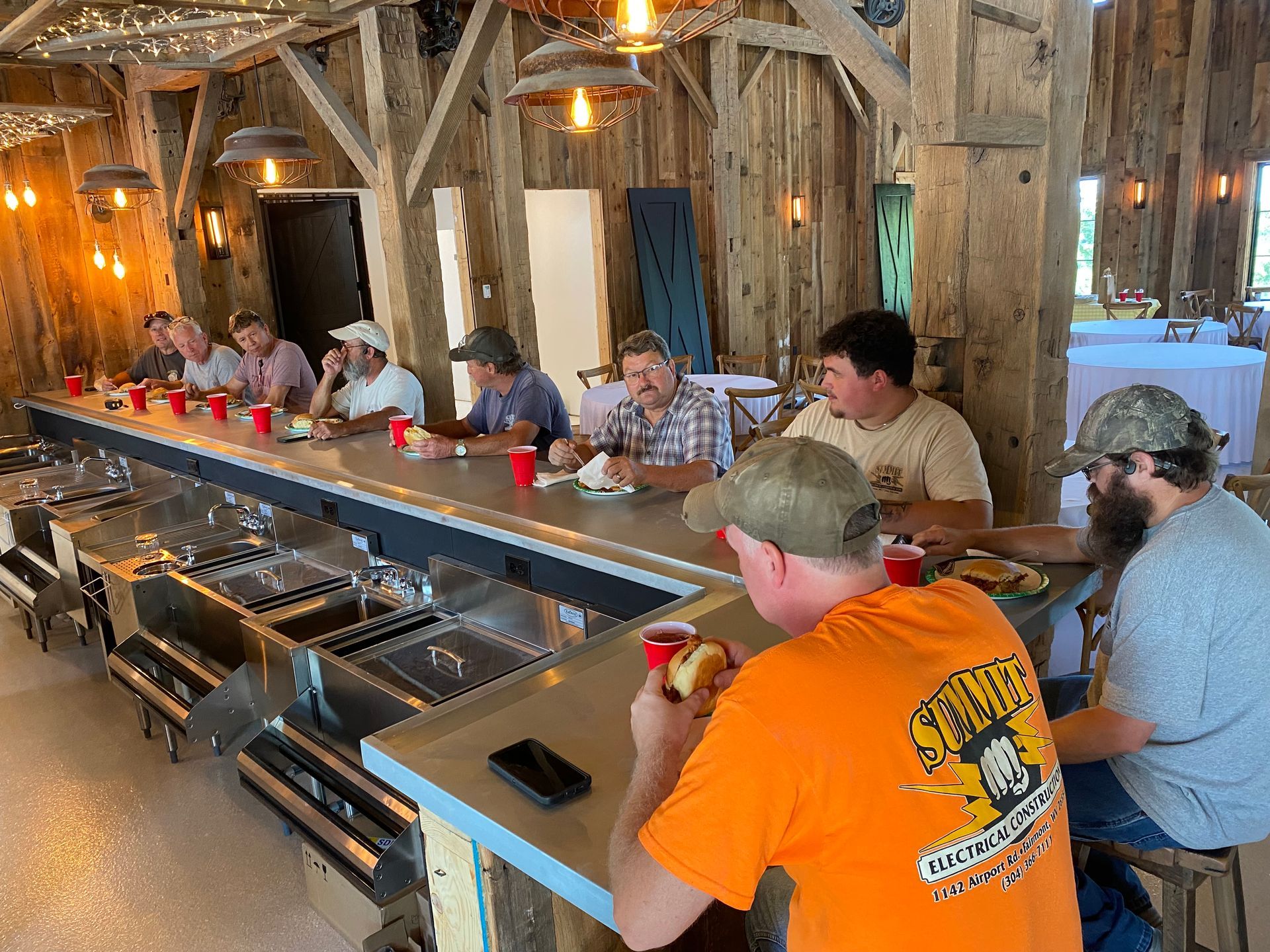 People eating at a long bar inside a wooden barn. They are eating and drinking, lit by hanging lights.