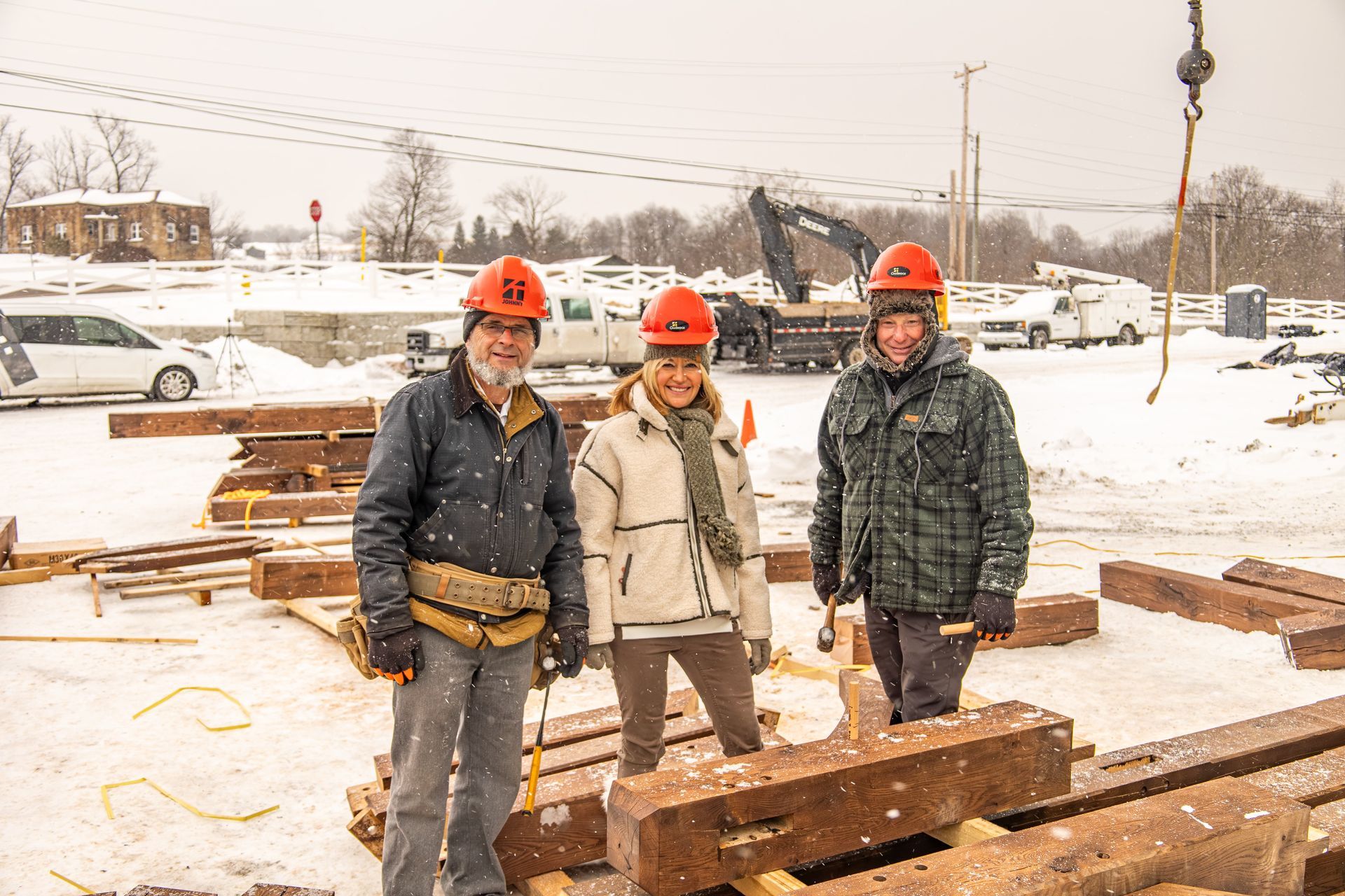 Three people in orange hard hats pose amidst construction beams in snowy conditions.