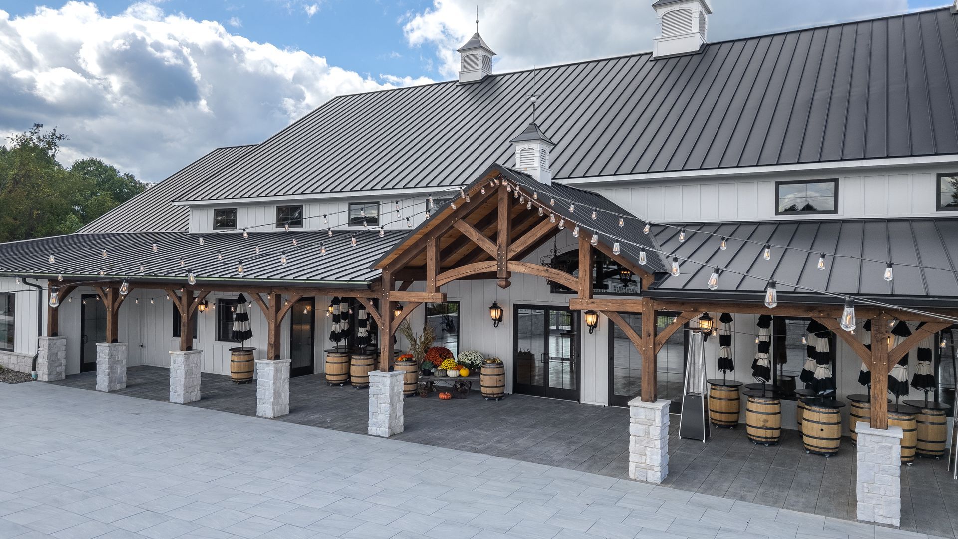 White barn with black roof, wooden porch, barrels, string lights, gray gravel.