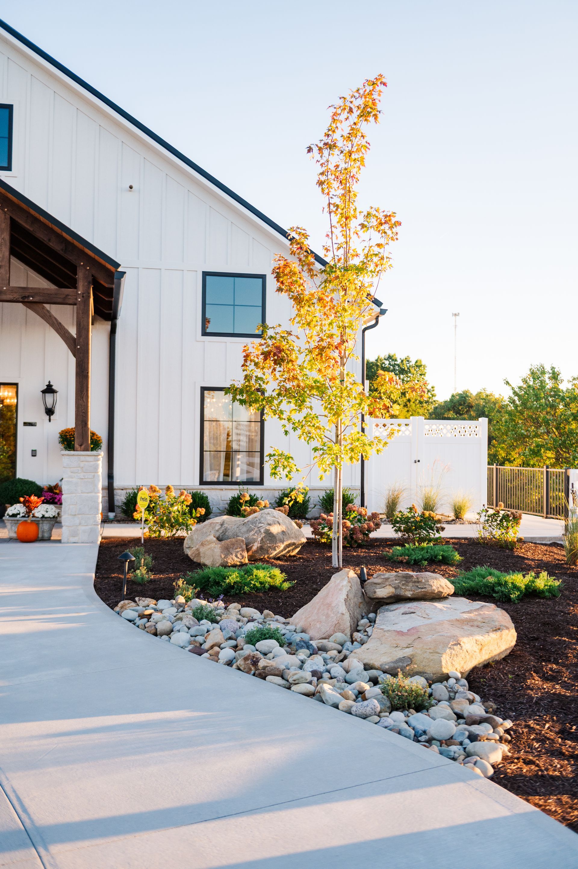 White ak 1938 with a modern entrance, stone and rock landscaping, and a young tree in the front.