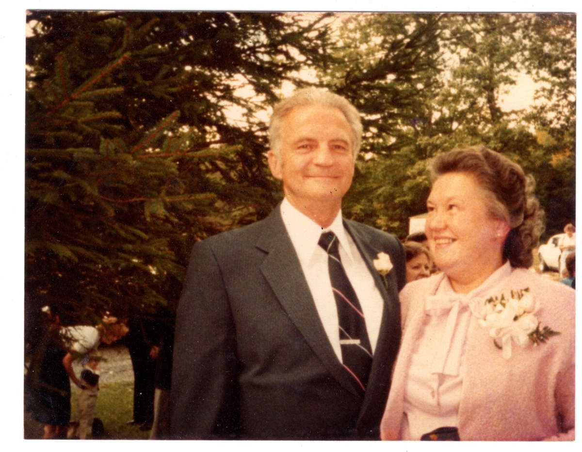 Man and woman, formally dressed, smile outside. Man wears a suit, woman, a pink suit.
