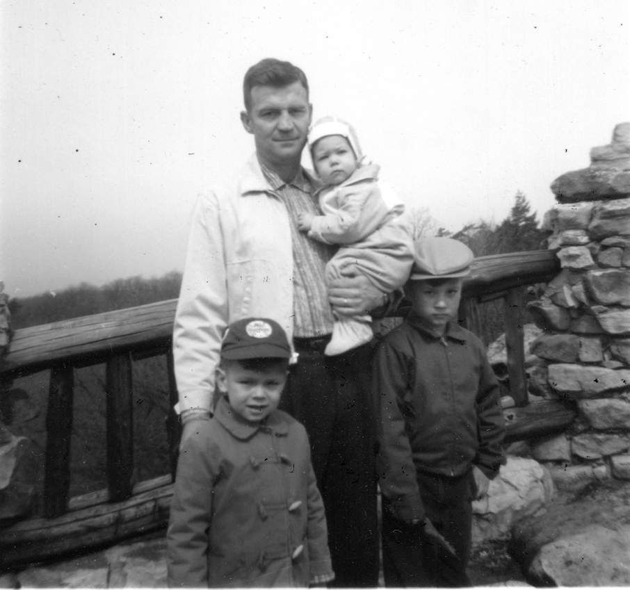 Man holding baby, standing with two young boys on a stone structure overlooking a wooded landscape.