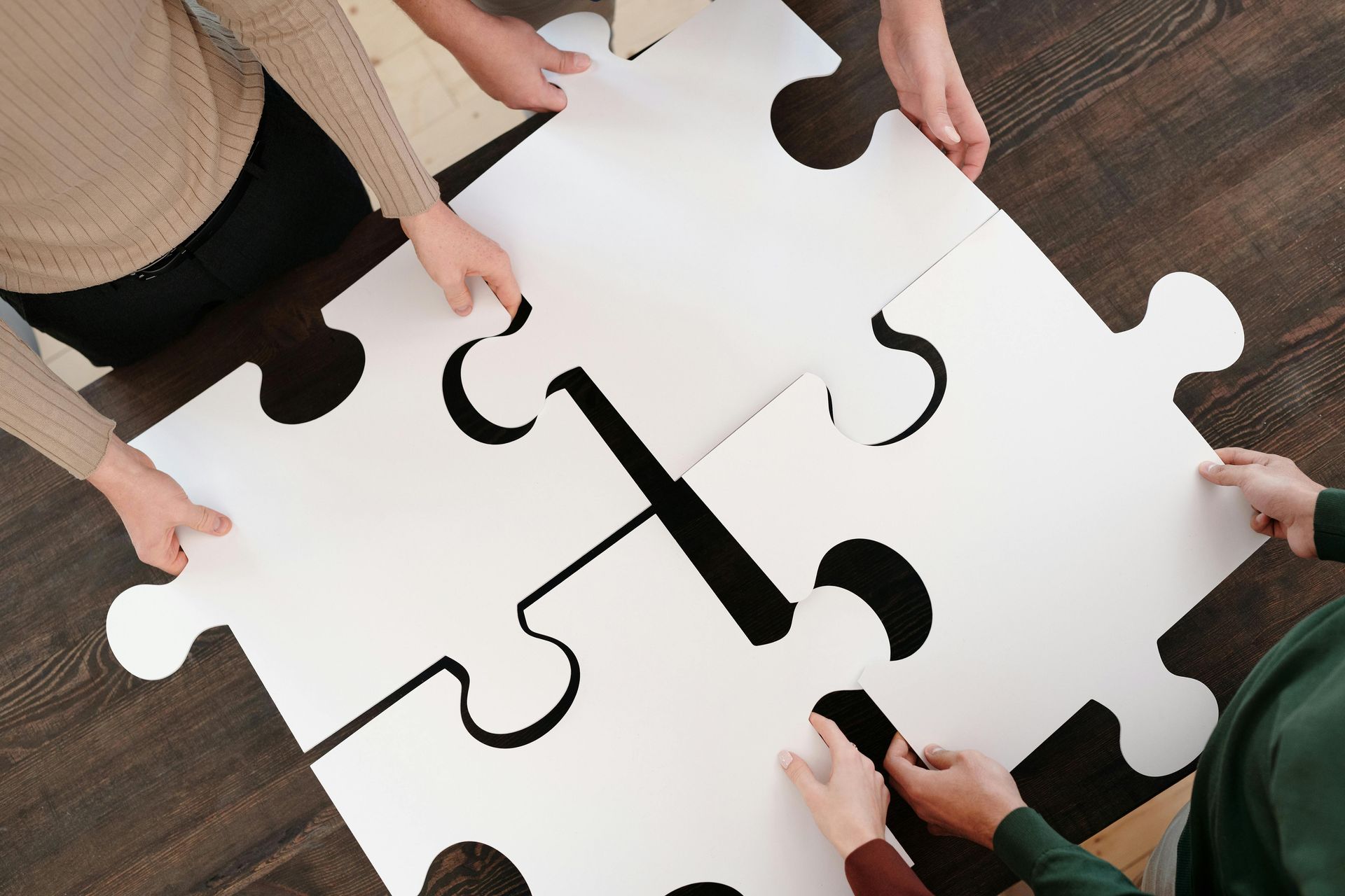 Vier Hände fügen vier schlichte weiße Puzzleteile auf einer dunklen Holzoberfläche zusammen.