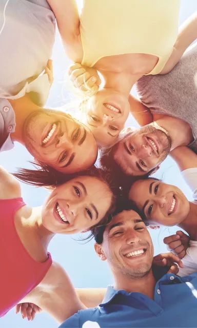 Eine Gruppe von Menschen steht im Kreis, die Köpfe zur Kamera geneigt, und lächelt unter einem strahlend blauen Himmel.